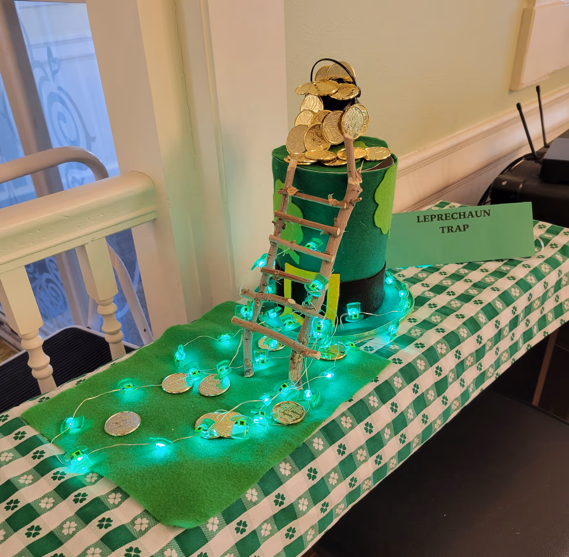 A decorative leprechaun trap setup on a green and white checkered tablecloth with shamrock patterns. The trap features a green felt hat with gold chocolate coins spilling out of the top, a small wooden ladder leaning against the hat, and green string lights surrounding the base. A sign next to the display reads 'LEPRECHAUN TRAP'.