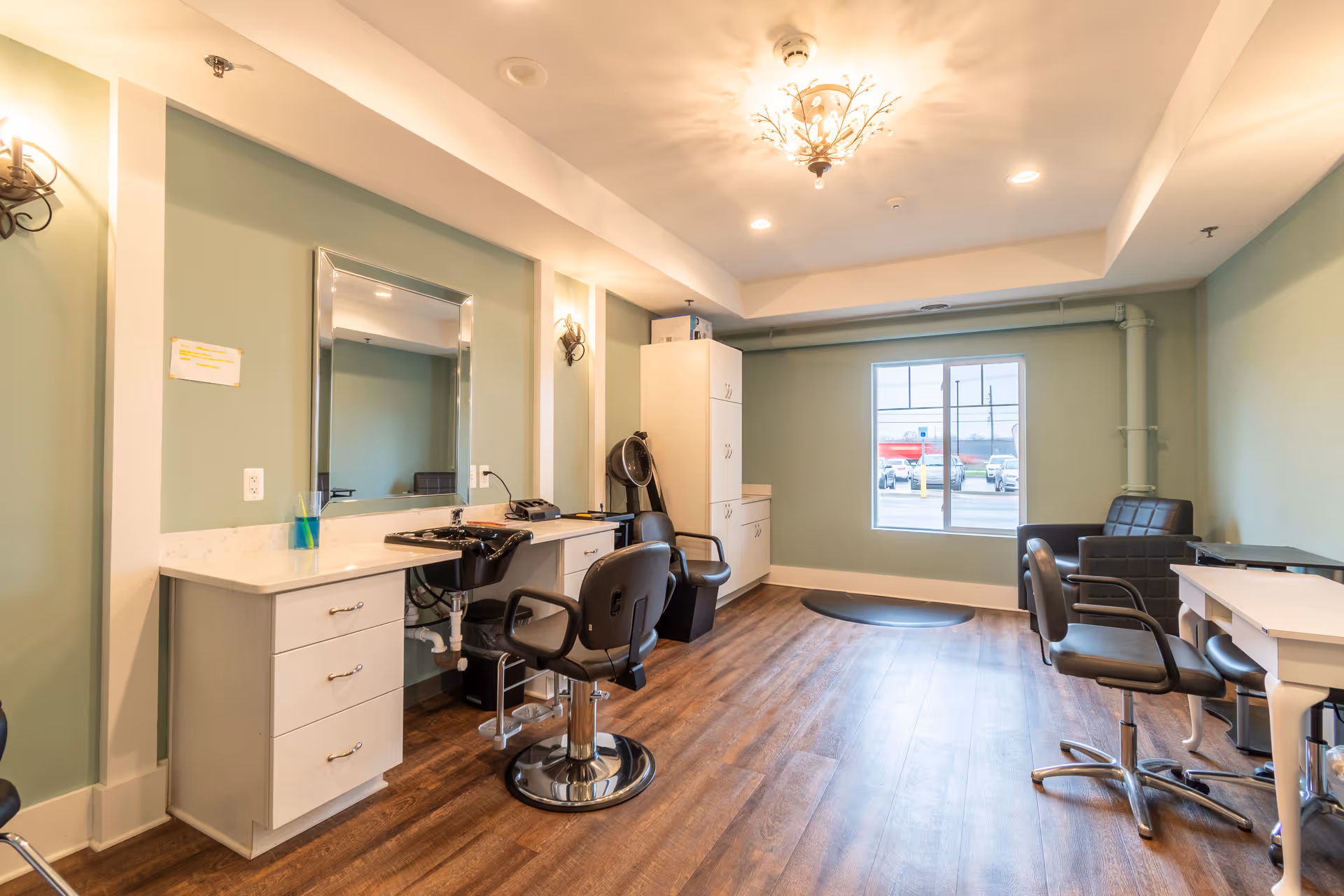 A bright and clean salon room with light green walls and wood flooring. The room features a styling station with a large mirror, a black salon chair, a white countertop with drawers, and a hair washing sink. There are additional black chairs and a small white table on the right side, and a window at the far end letting in natural light. The ceiling has recessed lighting and a decorative light fixture.