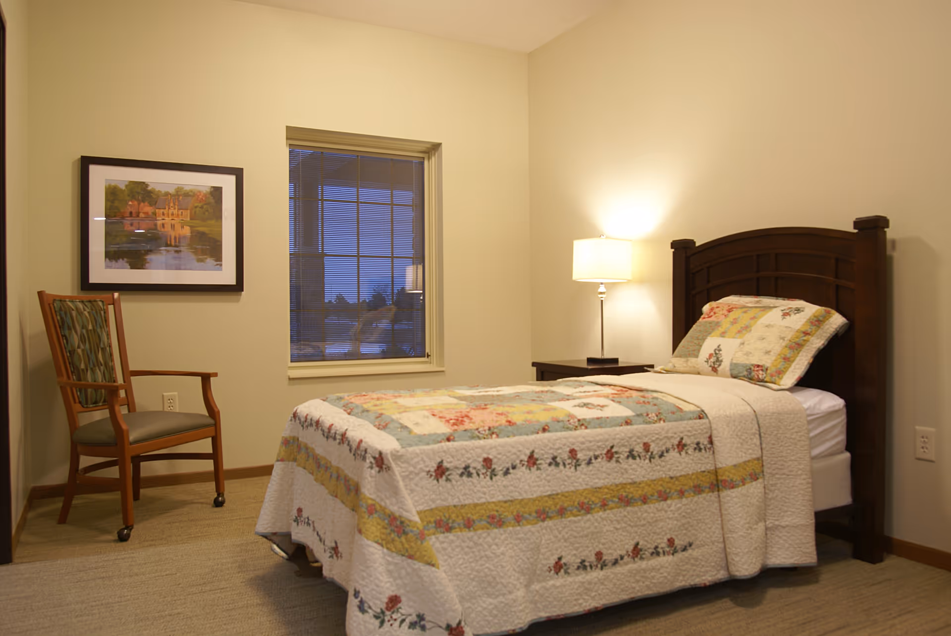 A cozy bedroom with a single bed featuring a floral patchwork quilt and matching pillow. Next to the bed is a wooden nightstand with a lit lamp. A wooden chair with patterned upholstery is placed near a window with blinds. A framed painting of a house by a lake hangs on the wall.