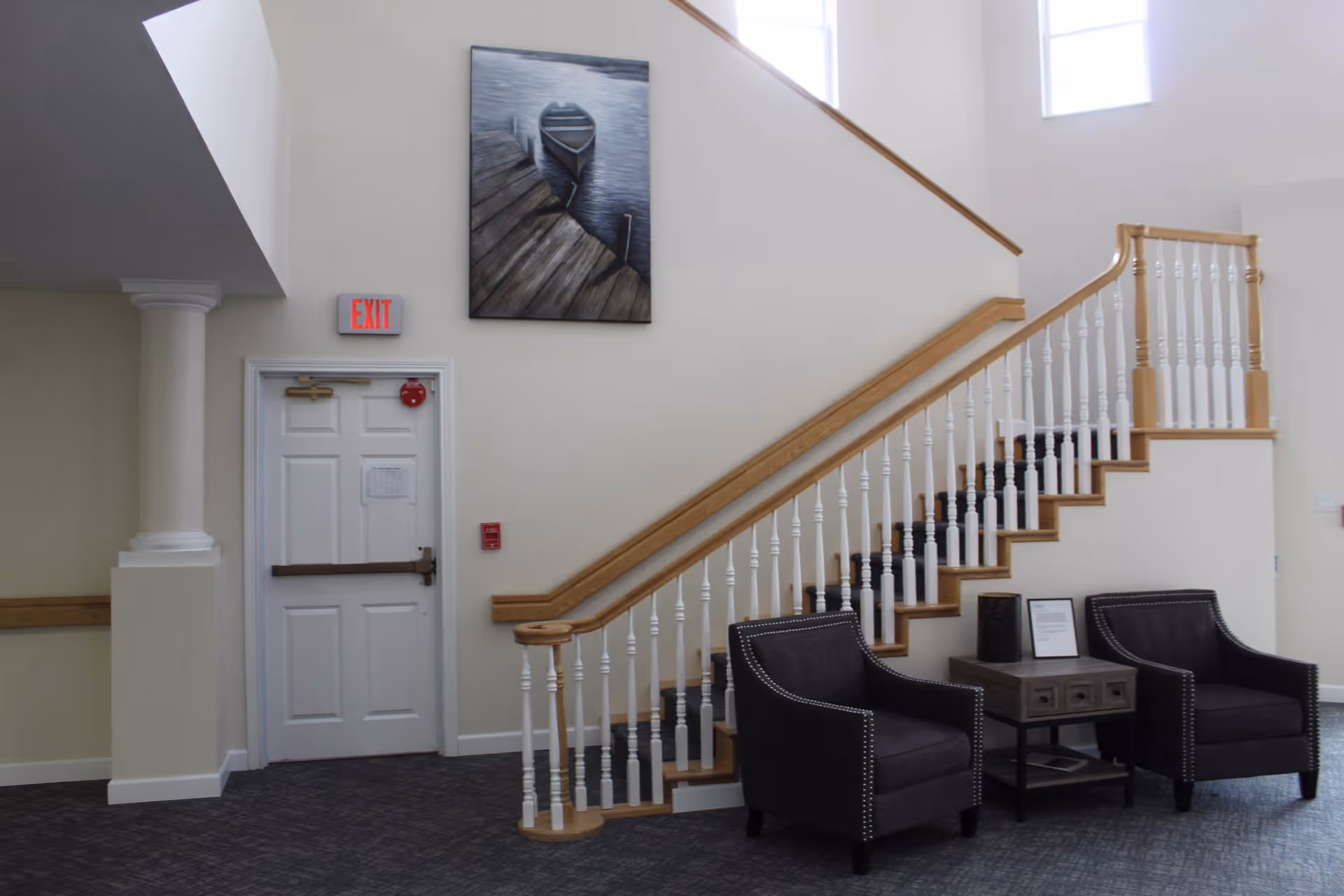 Interior view of a senior living facility showing a staircase with wooden handrails and white balusters. Two black armchairs with nailhead trim are placed on either side of a small wooden table with drawers. Above the door next to the staircase is an exit sign, and a painting of a boat docked at a pier hangs on the wall. The area is well-lit with natural light coming from a window above.
