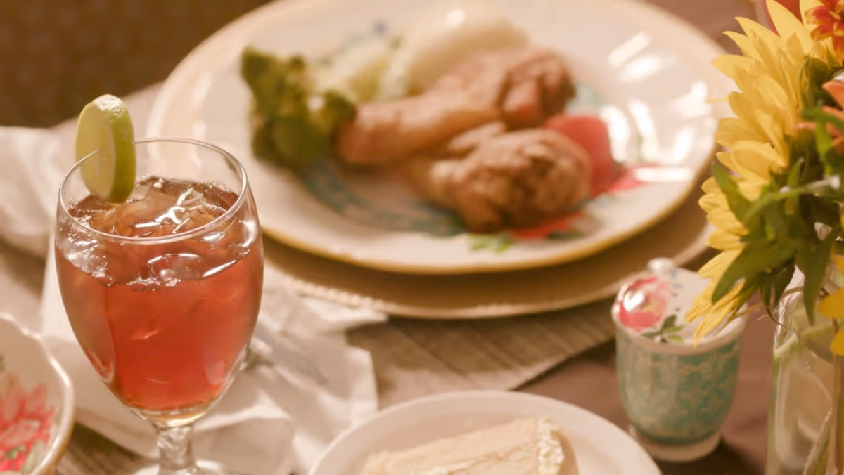 A close-up view of a dining table set with a glass of iced tea garnished with a lime wedge, a plate with two pieces of fried chicken and some broccoli, a small decorative container, and a vase with yellow flowers.