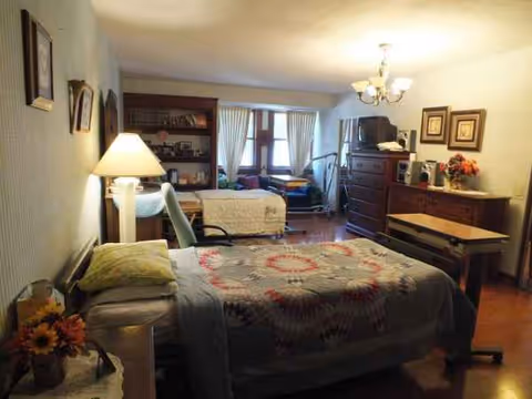 Cozy senior living bedroom with a quilted bed, nightstand, lamp, dresser, and seating.