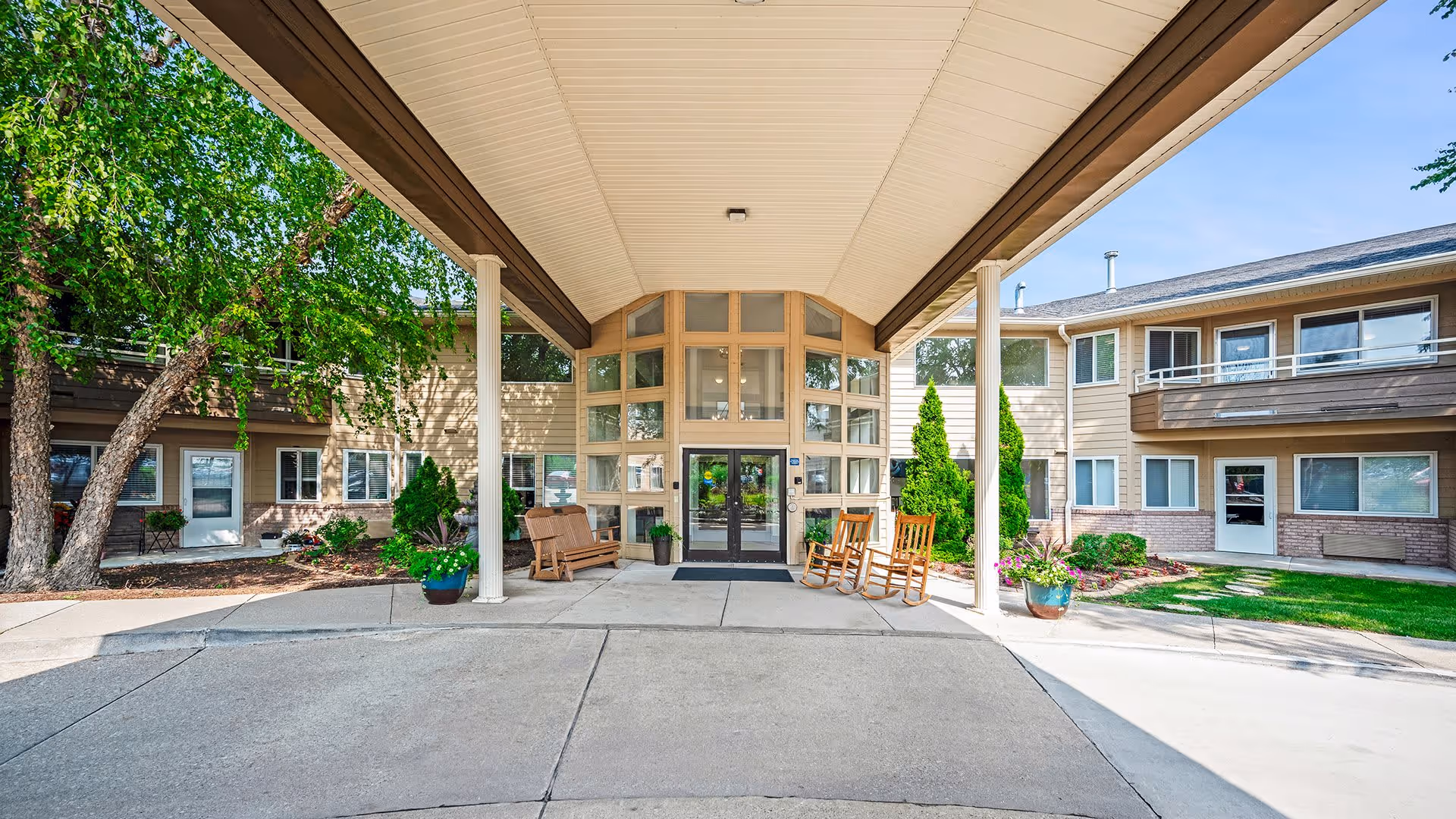 Entrance of a senior living facility with a covered driveway, two rocking chairs, a bench, potted plants, and a two-story building with multiple windows and balconies under a clear blue sky.