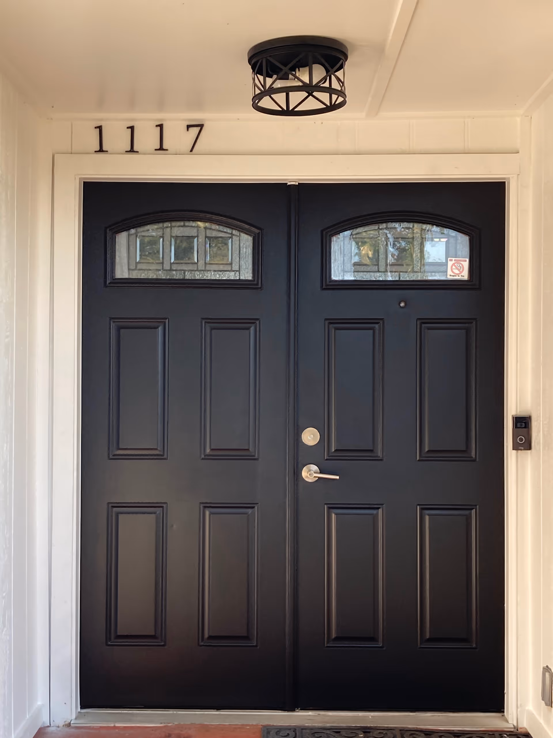 Double black front doors with decorative glass panels at the top, a silver door handle and lock on the right door, a black ceiling light fixture above, and the house number 1117 displayed above the doors.