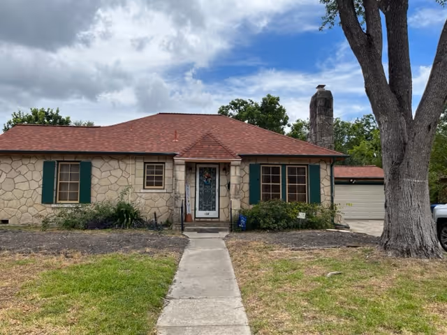 Single-story stone house with a red roof, central front door, green shutters, a concrete walkway, and a large tree in the front yard.