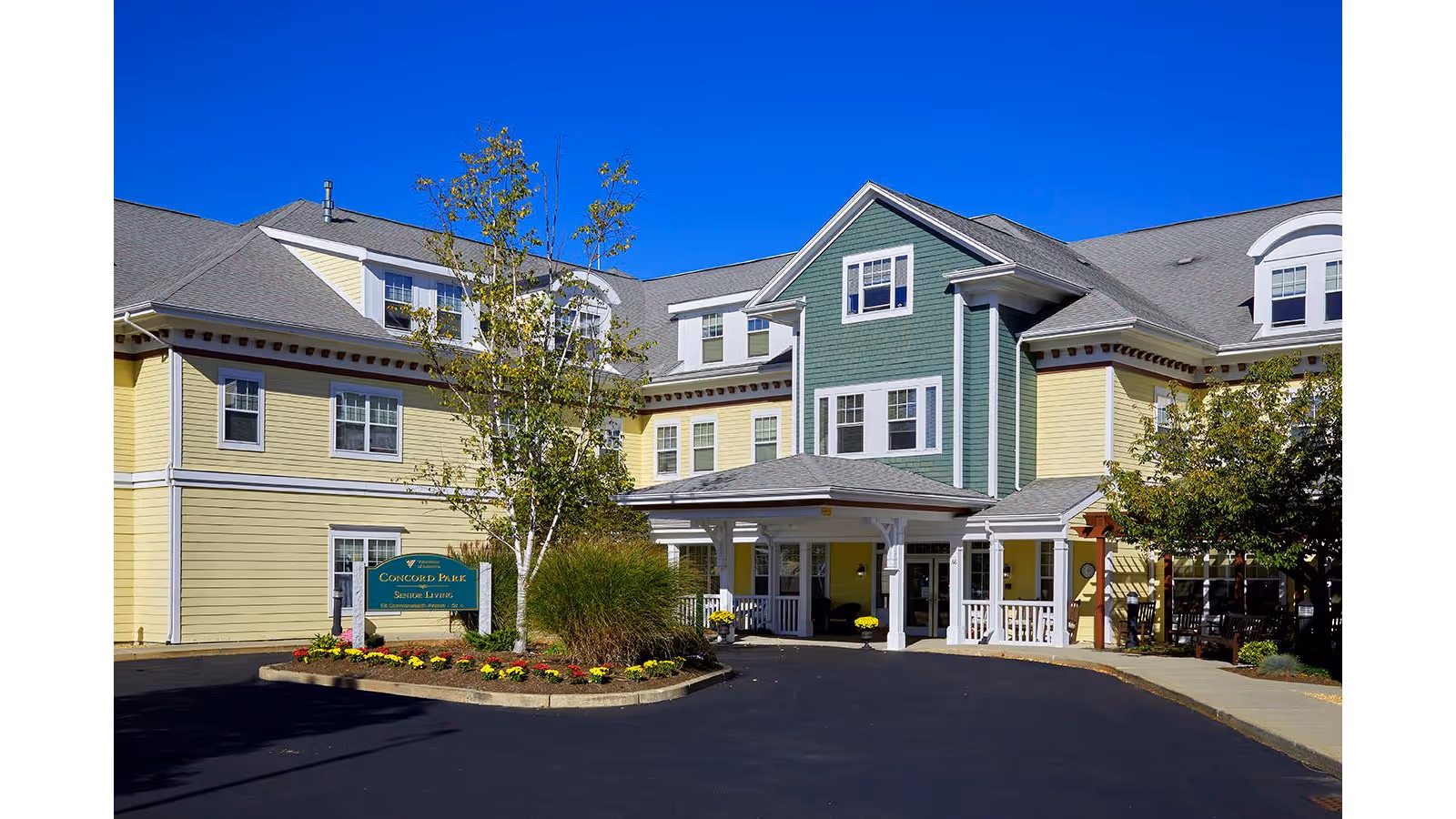 Exterior view of Concord Park Senior Living facility showing a multi-story building with yellow and green siding, multiple windows, a covered entrance, landscaped flower beds, and a clear blue sky.