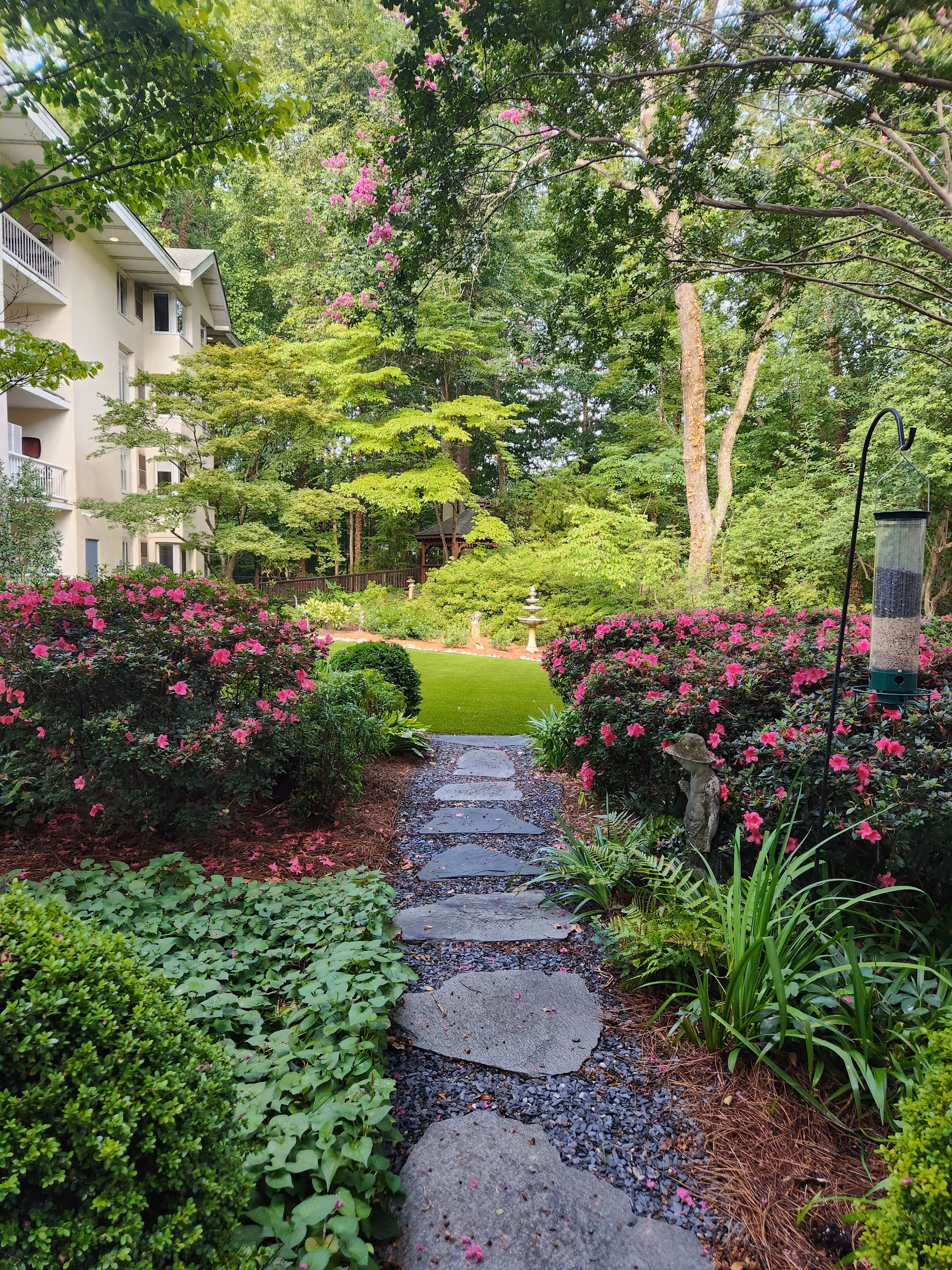 A peaceful garden path made of stepping stones surrounded by lush greenery and blooming pink flowers. To the left, part of a multi-story building with balconies is visible. The garden features various plants, trees, and a bird feeder on the right side of the path.