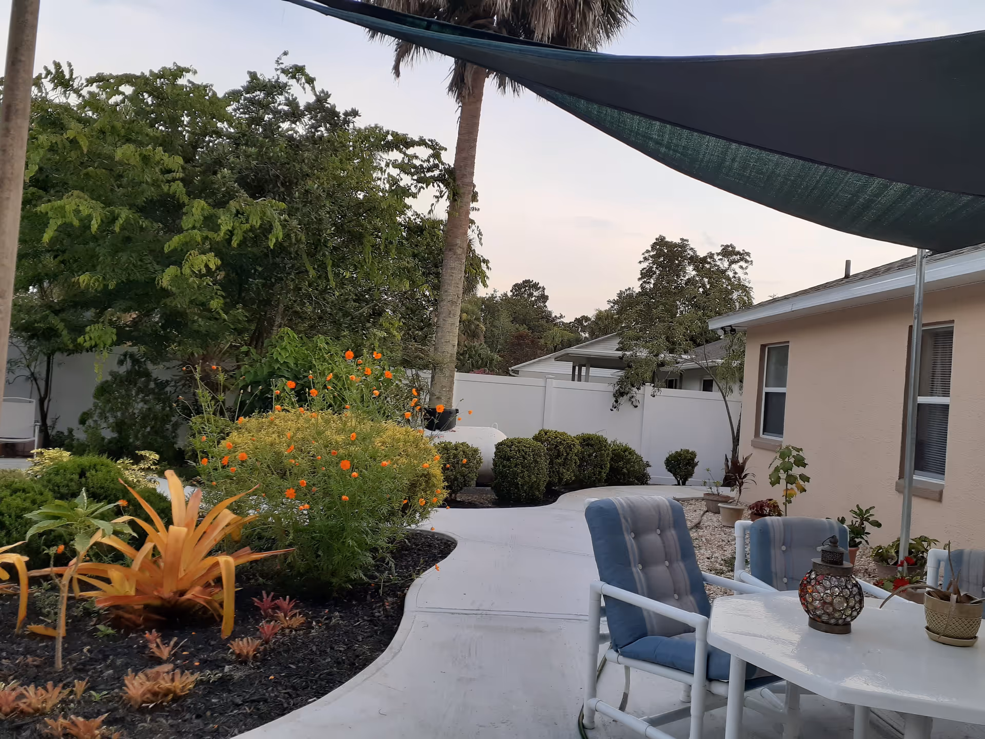 Outdoor patio area with a white curved walkway surrounded by various green plants and bushes, including some orange flowers. There is a table with blue cushioned chairs under a dark shade sail attached to poles. A beige building with windows is visible on the right side.
