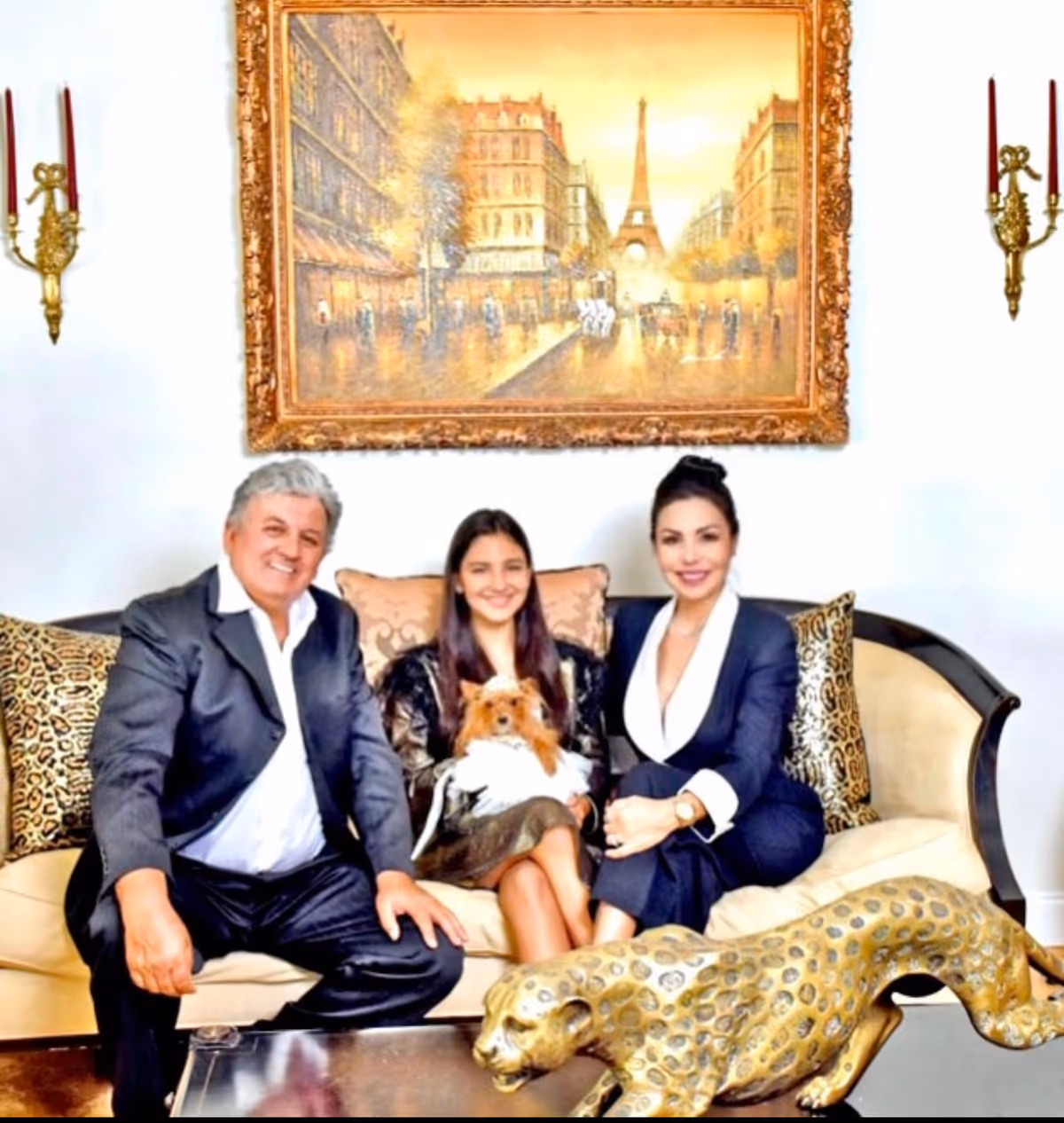 Three people seated on an upholstered sofa holding a small dog in a decorated living room with a large framed painting, wall sconces, and a decorative leopard sculpture on the coffee table.