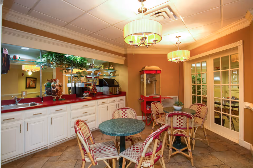Warmly lit common dining area with round tables and wicker chairs, a red countertop with appliances and a popcorn machine.