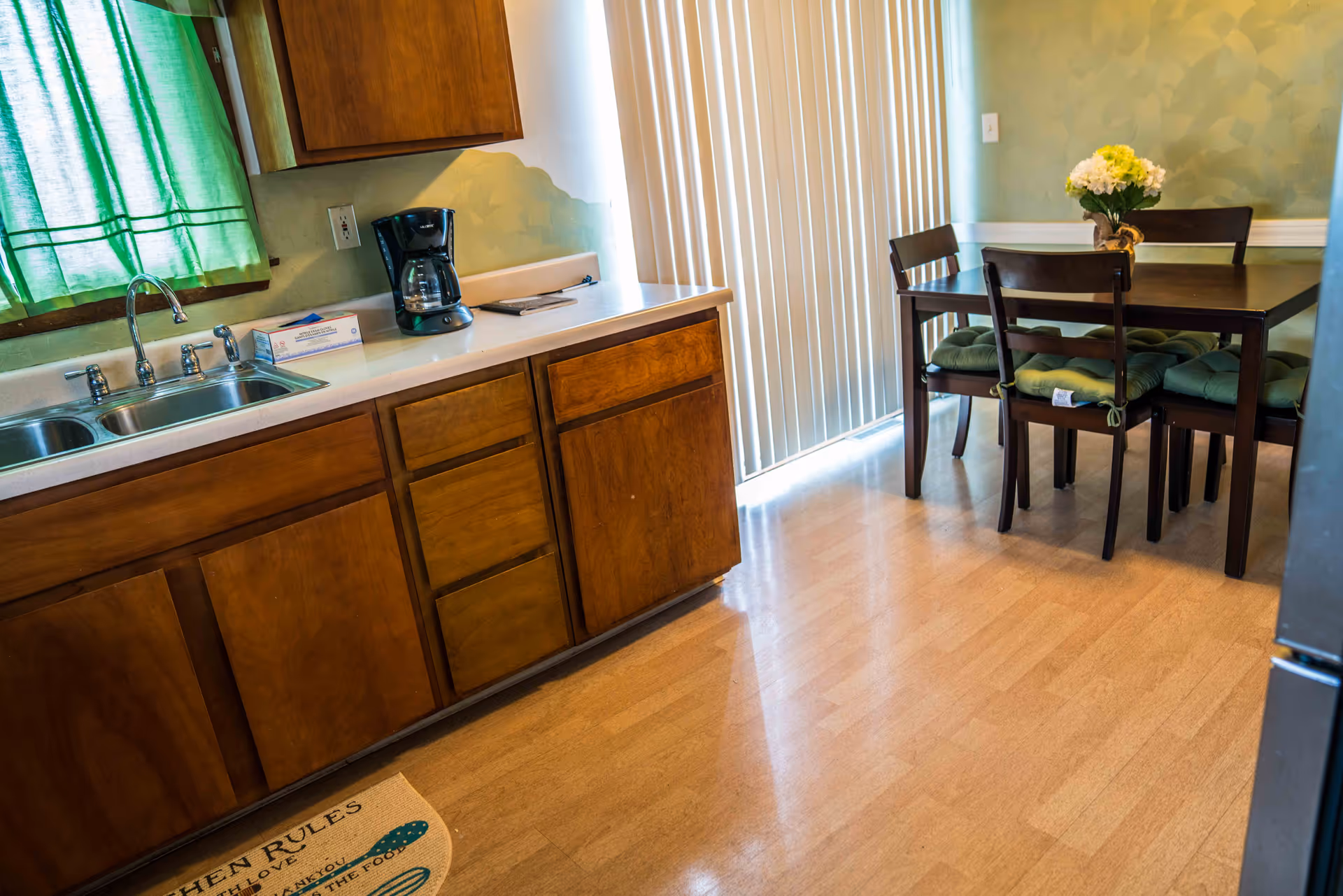 A kitchen and dining area with wooden cabinets, a double sink, a coffee maker on the counter, and a dining table with four chairs that have green cushions. There is a window with green curtains and vertical blinds covering a sliding door. A small flower arrangement is on the dining table.
