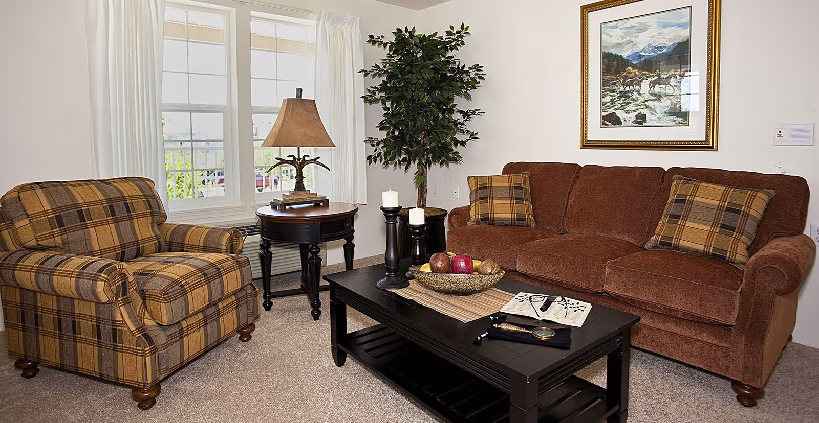 A cozy living room with a brown sofa and a plaid armchair. There is a black coffee table with decorative candles, a bowl of fruit, and a newspaper with reading glasses on it. A round side table with a lamp is placed next to the armchair. A large window with white curtains lets in natural light, and a framed painting of a mountain scene hangs on the wall above the sofa. A tall potted plant is in the corner.
