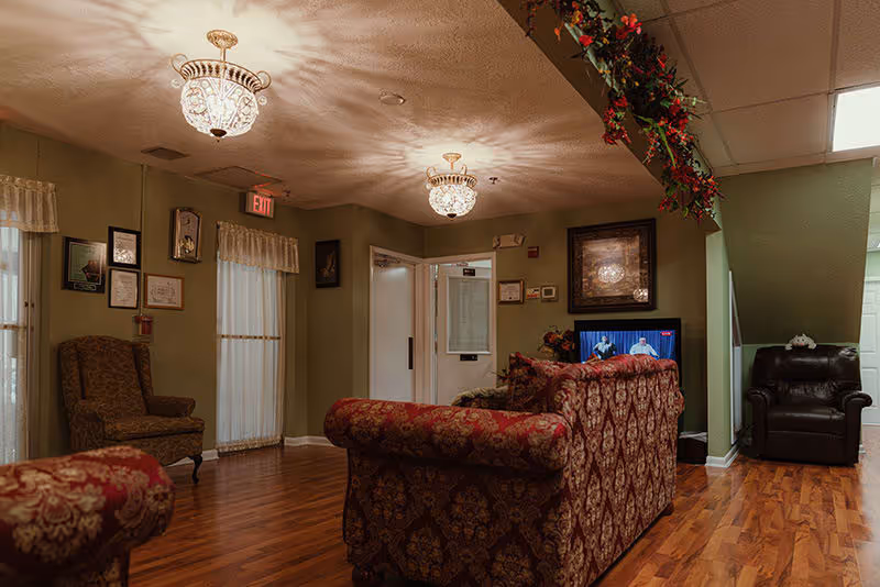 A cozy living room area in an assisted living facility with patterned upholstered sofas and armchairs, wooden flooring, decorative ceiling lights, framed pictures on the walls, and a television showing a program. There is a garland decoration hanging from the ceiling near the corner.