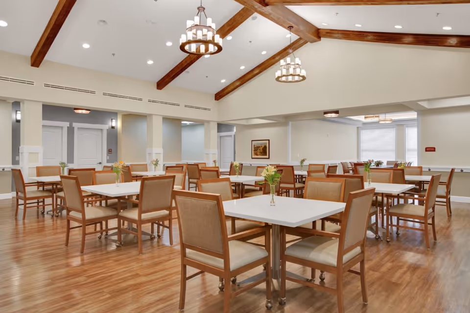 Spacious dining room with multiple tables and chairs, exposed wooden ceiling beams, chandeliers, and vases of flowers on the tables.