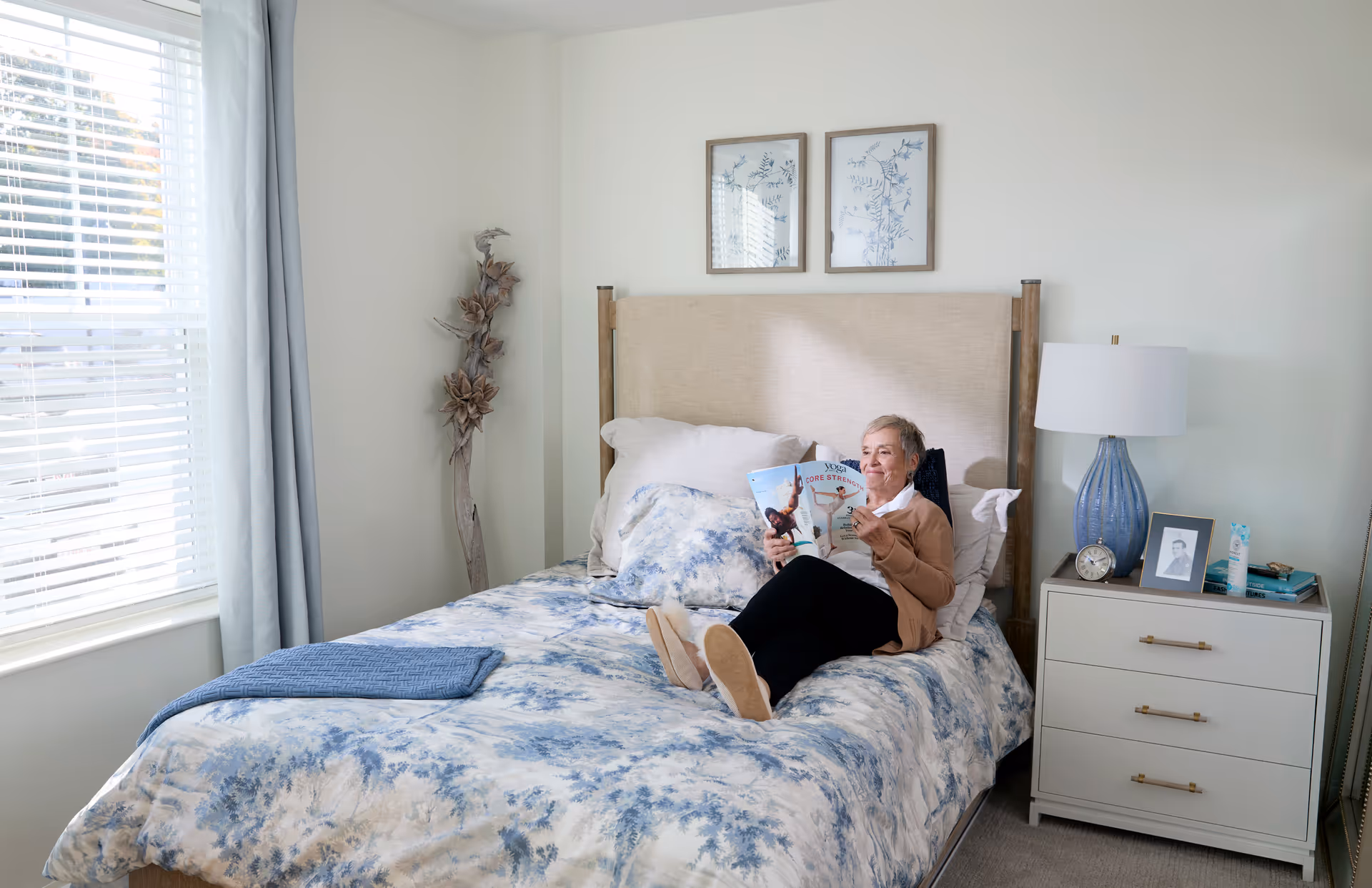 A senior woman relaxing on a bed in a bright bedroom, reading a magazine. The room features a large window with white blinds and light blue curtains, a beige upholstered headboard, blue and white bedding, a nightstand with a blue lamp, a clock, a framed photo, and some books. Two framed botanical prints hang on the wall above the bed.