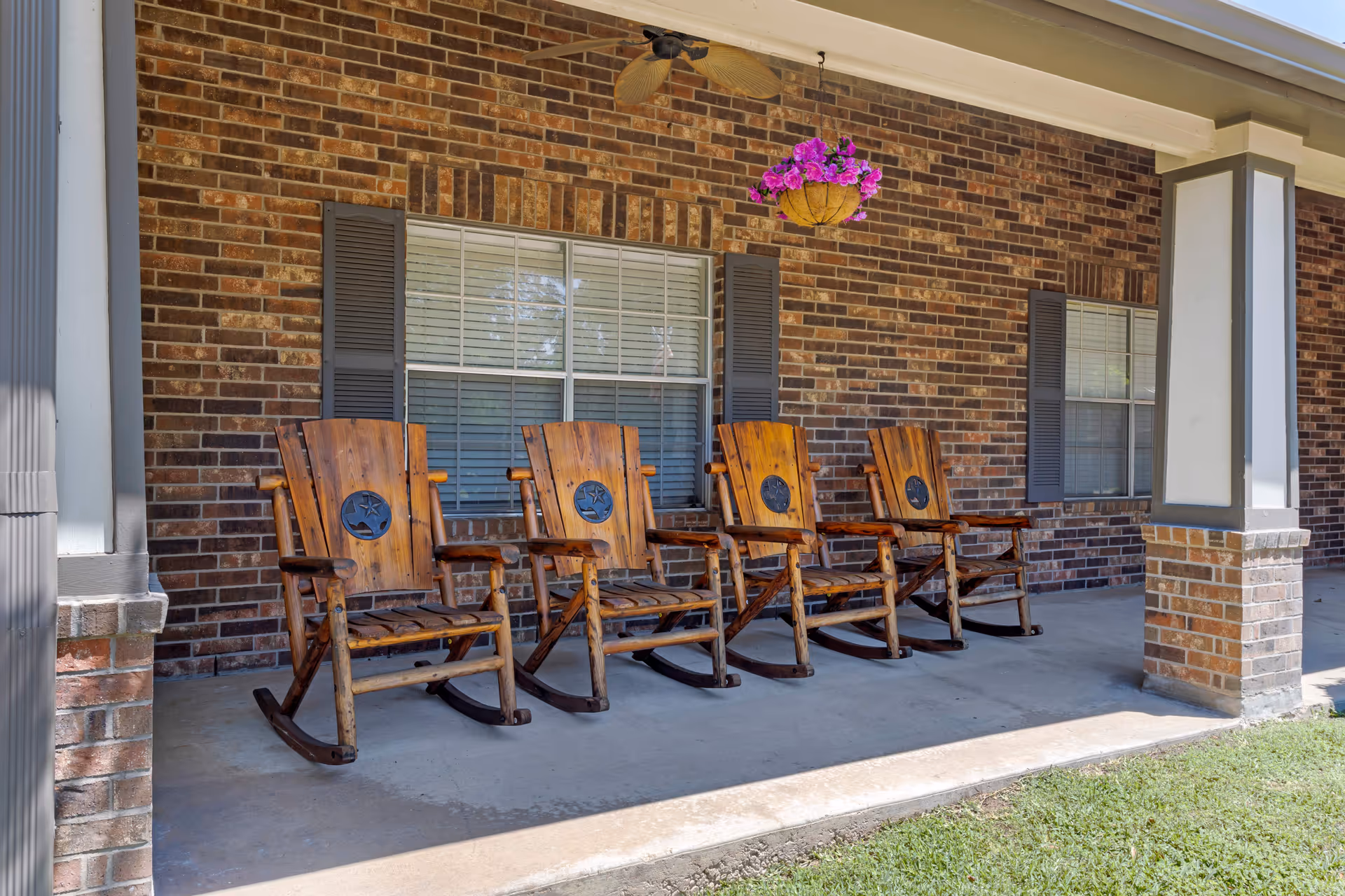 A covered outdoor porch area with four wooden rocking chairs lined up in front of a brick wall with two windows. A hanging basket with purple flowers is suspended from the ceiling, and a ceiling fan is mounted above the chairs. The porch has concrete flooring and is supported by brick and white pillars.