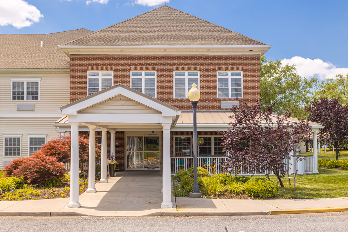 Front entrance of a two-story brick and siding senior living building with a covered porte-cochère, porch, lamppost, and landscaped shrubs.
