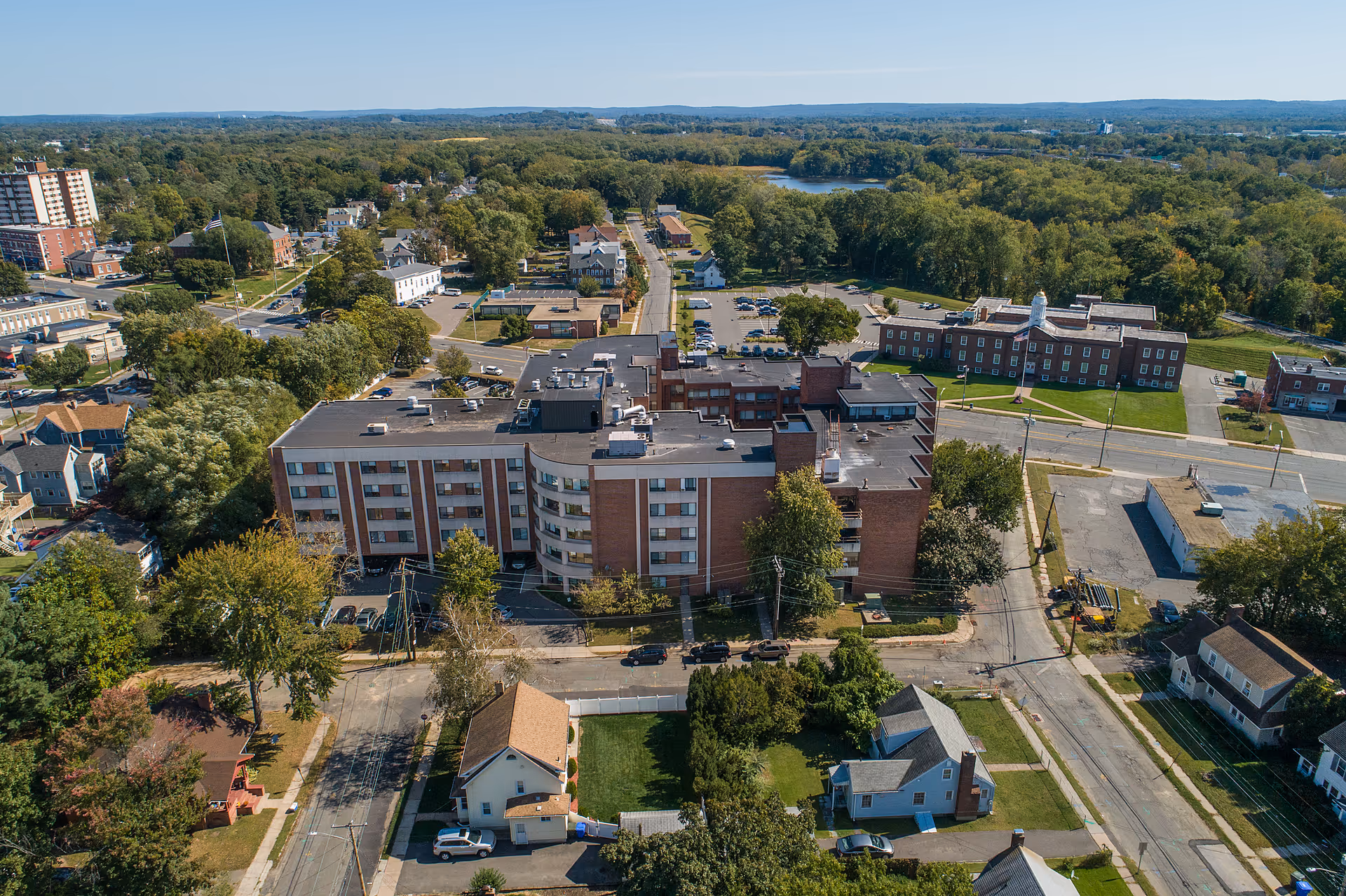 Aerial view of Riverside Health and Rehabilitation Center, a multi-story brick building surrounded by trees, roads, and smaller houses in a suburban area with a river and forested area in the background.