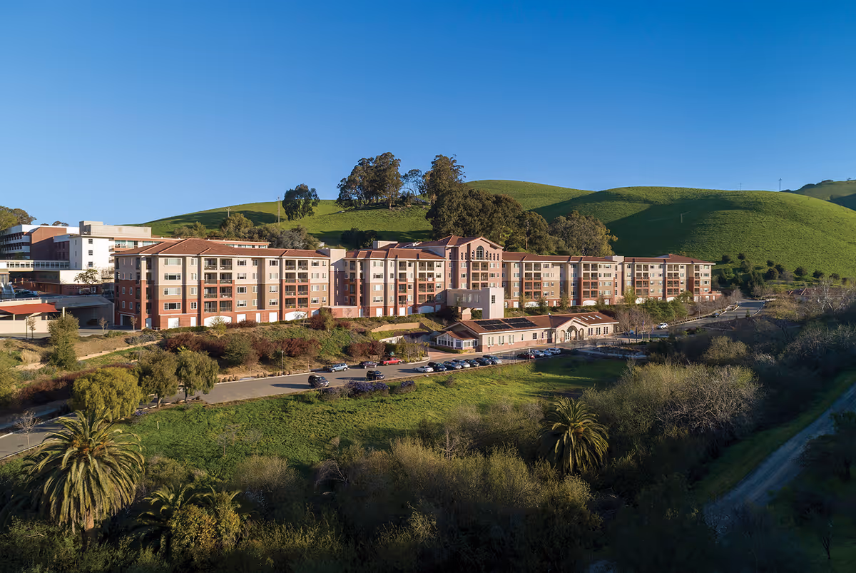 A large multi-story retirement community building with a red and beige exterior, situated in a green hilly landscape under a clear blue sky. There are several cars parked in front of the building and trees and shrubs surrounding the area.