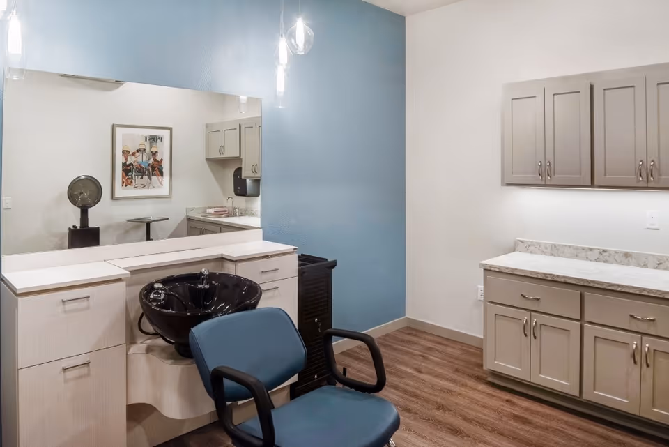 A salon-style hair-washing station with a black basin, blue chair, large mirror, and surrounding cabinets in a bright room.