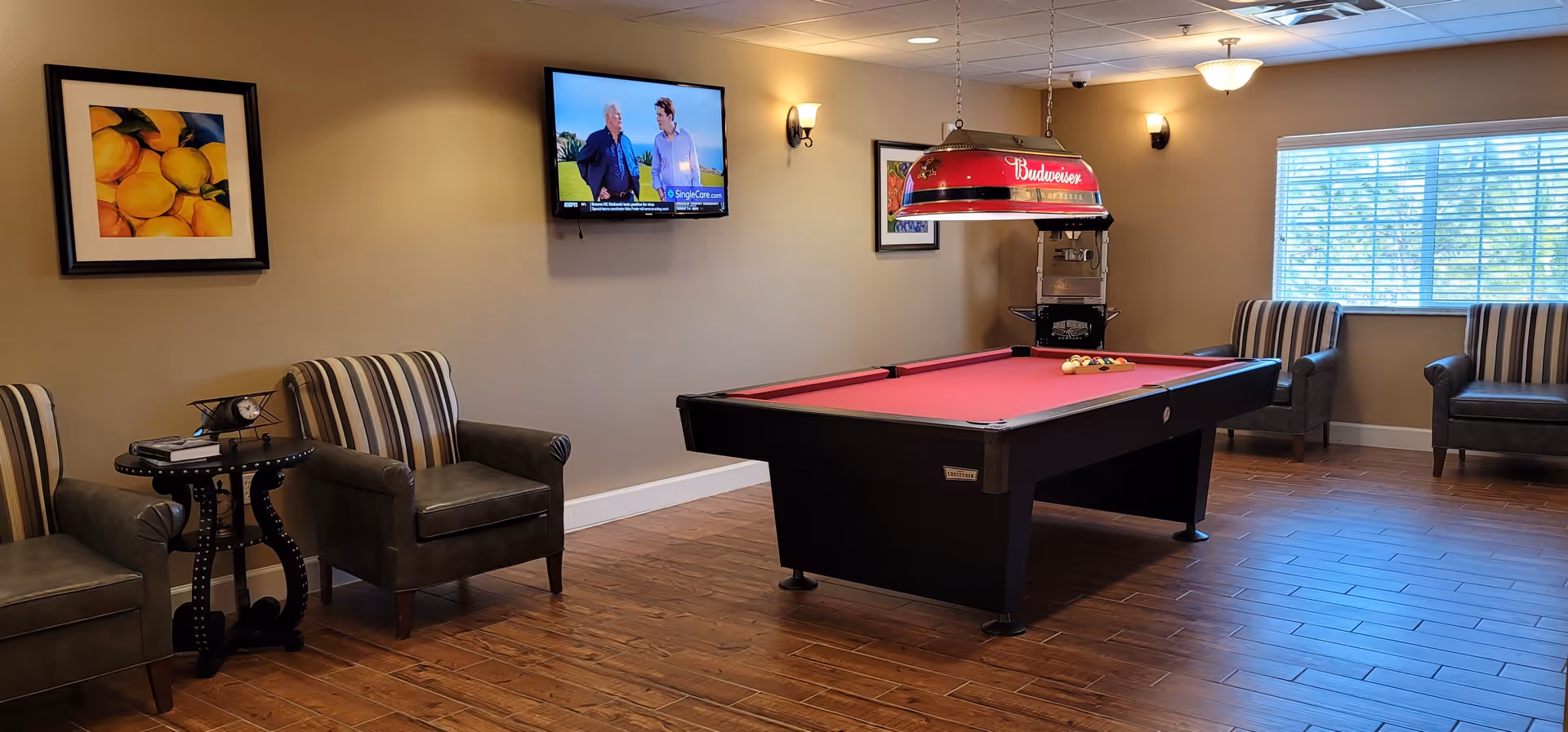 A recreational room with a red felt pool table in the center, a Budweiser-branded hanging light above it, and a popcorn machine in the corner. The room has wooden flooring, beige walls, a wall-mounted TV showing two people, and several striped and leather armchairs arranged along the walls. Two framed pictures hang on the walls, and a window with blinds lets in natural light.