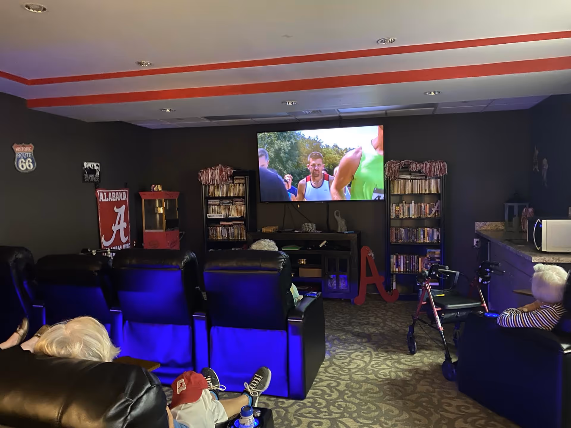 A cozy entertainment room with several black leather recliners facing a wall-mounted TV showing a sports event. The room has dark walls with red trim near the ceiling, bookshelves filled with DVDs or books, a popcorn machine, and a microwave on a counter. Two elderly individuals are seated in the recliners, one with a walker nearby. There are Alabama-themed decorations on the walls and floor.