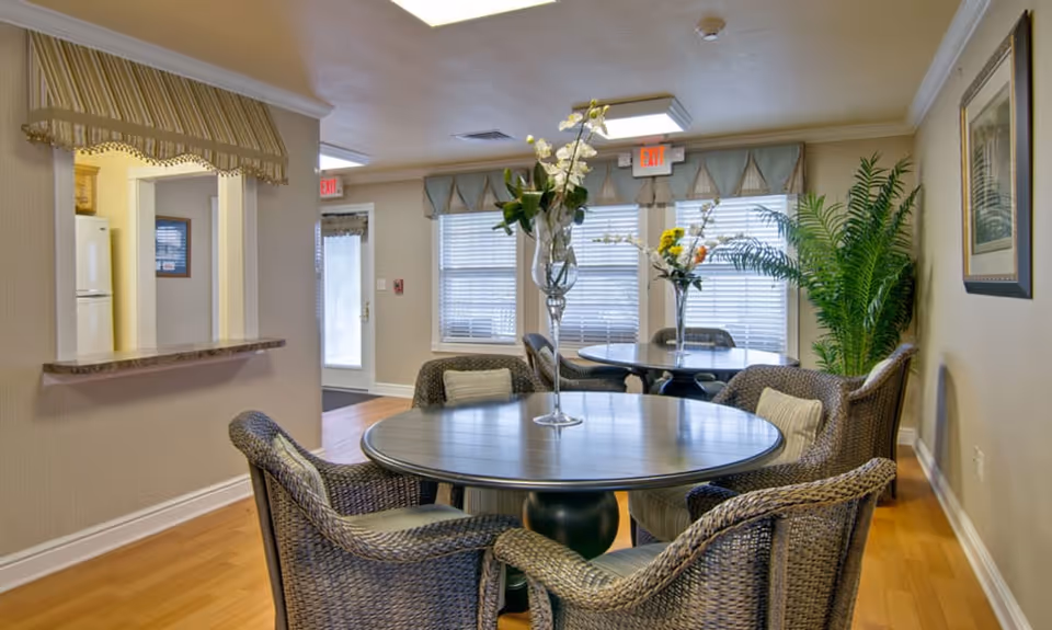 A cozy dining area with two round tables, each surrounded by four wicker chairs with cushions. Each table has a tall vase with flowers. The room has light-colored walls, wood flooring, and large windows with blinds and valances. There is a pass-through window to a kitchen area on the left and a large green plant on the right. Exit signs are visible above the doors in the background.