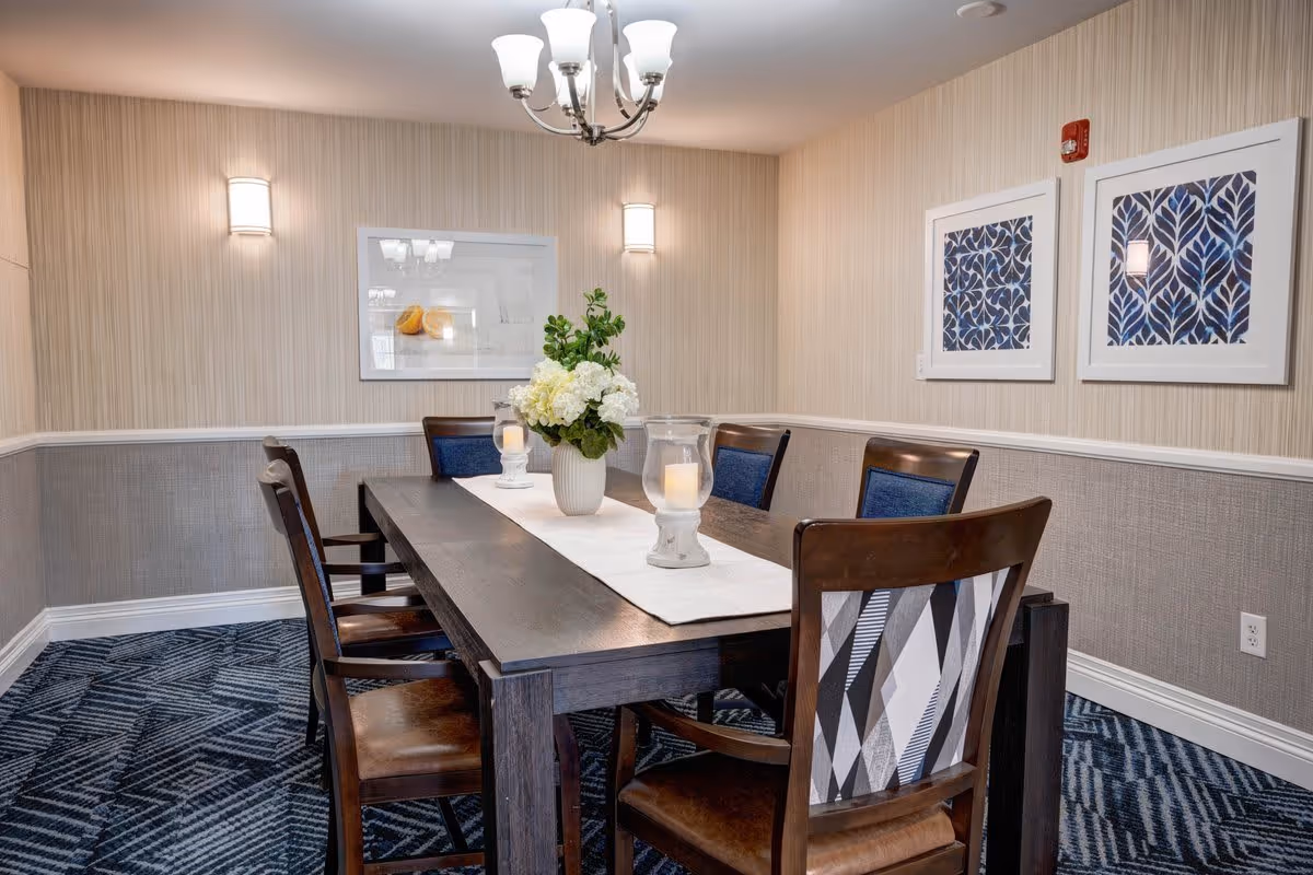 A dining room with a dark wooden table surrounded by six chairs, some with patterned backs and others with blue upholstery. The table is decorated with a white runner, a vase of white flowers, and two candle holders. The room has patterned blue carpet, beige and gray textured walls, three framed artworks, two wall sconces, and a chandelier overhead.