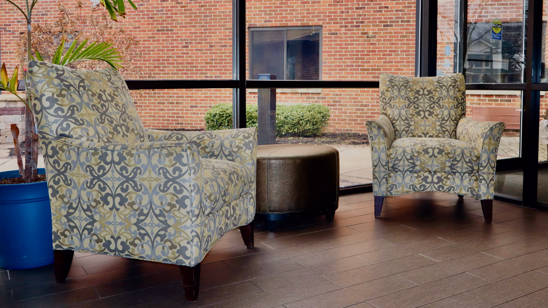 Two patterned armchairs with a small round ottoman between them, placed on a wooden floor in front of large glass windows showing a brick building and some greenery outside. A blue planter with a plant is visible to the left.