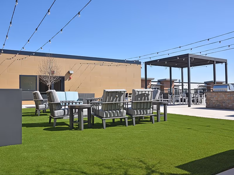 Outdoor seating area on a rooftop with cushioned chairs and tables arranged on green artificial grass under string lights, with a pergola and barbecue grill in the background against a clear blue sky.