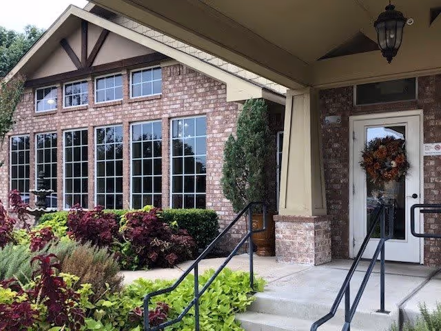 Entrance of a brick building with large windows and a covered porch. There are plants and shrubs along the walkway leading to the door, which has a decorative wreath hanging on it.