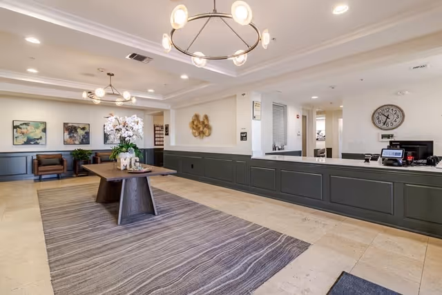 A spacious reception area with a long dark green counter on the right side, a large wooden table with a flower arrangement in the center on a striped rug, two brown leather chairs against the far wall, modern circular light fixtures on the ceiling, and decorative artwork on the walls.
