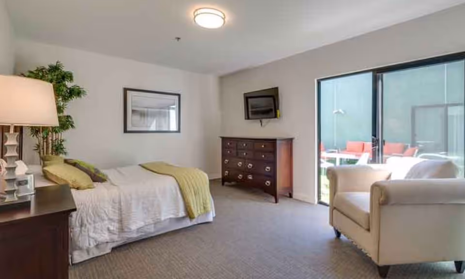 A senior living bedroom with a bed covered in white bedding and green pillows and throw. There is a wooden nightstand with a lamp and a plant next to the bed. Across from the bed is a wooden dresser with a flat-screen TV mounted above it. A beige armchair is positioned near a large sliding glass door that opens to an outdoor patio area with red cushioned seating.