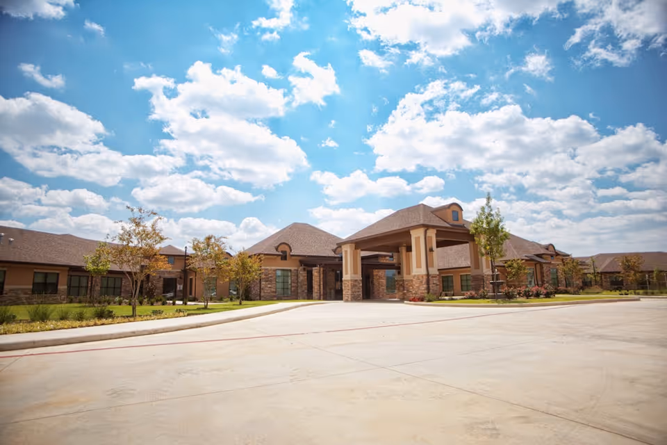 Single-story senior living facility entrance with a porte-cochere, landscaped grounds, and a blue sky with clouds.
