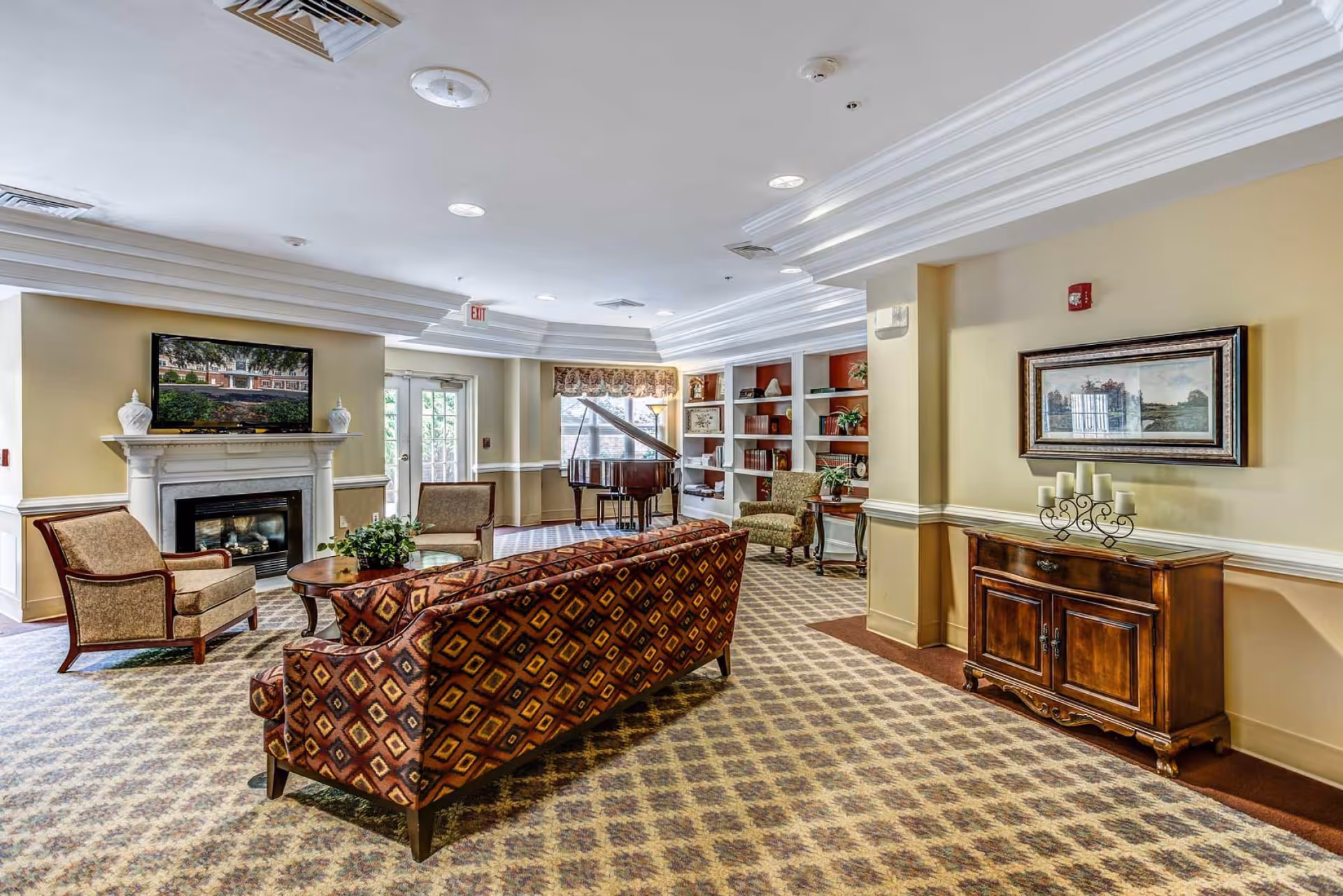 Communal living room with patterned sofa and chairs, a fireplace, piano, and built-in bookshelves.