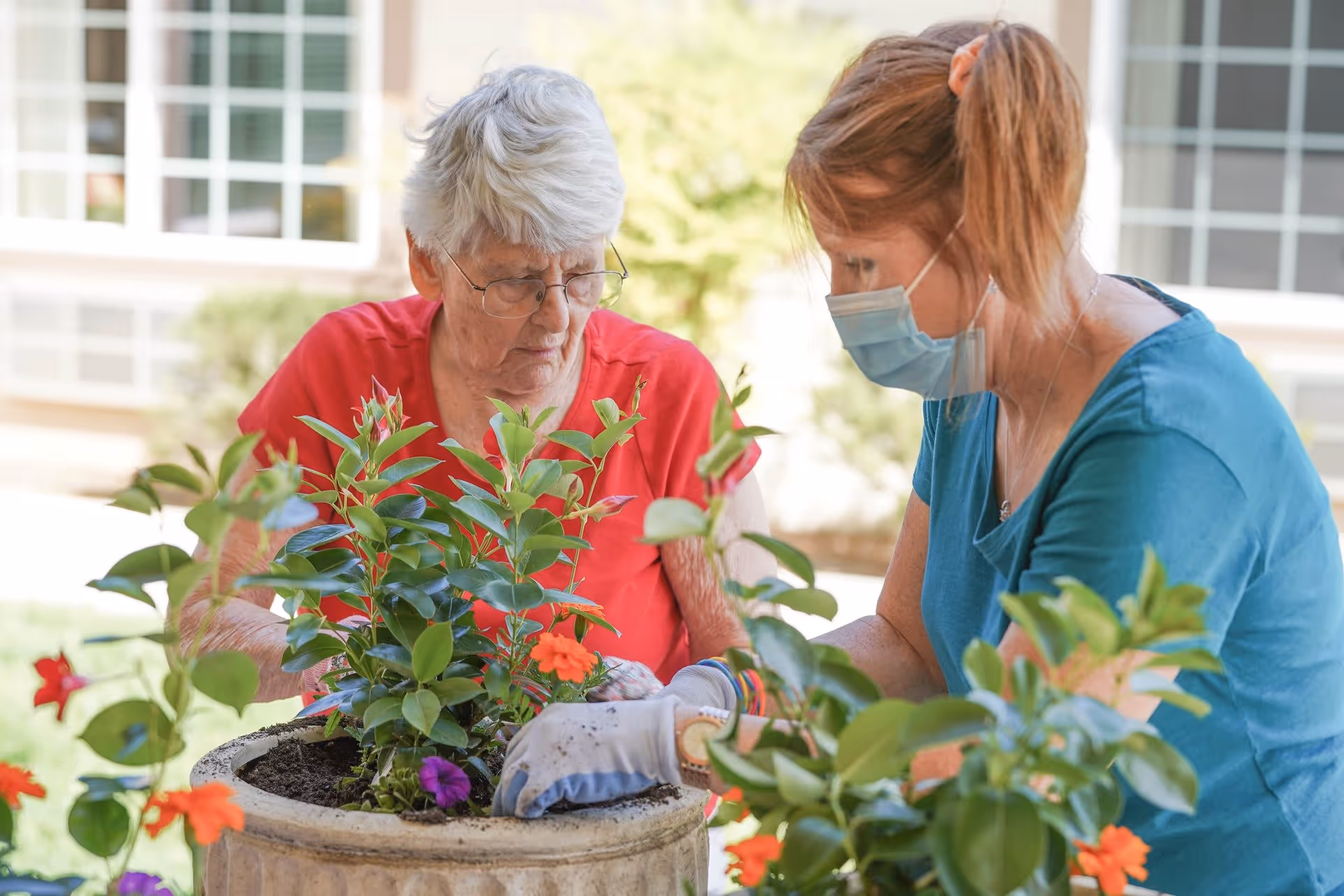 An elderly woman and a caregiver wearing a face mask are planting flowers together in a large outdoor planter. The elderly woman is wearing a red shirt and glasses, while the caregiver is wearing a blue shirt and gardening gloves. They are focused on arranging the plants, with a building visible in the background.