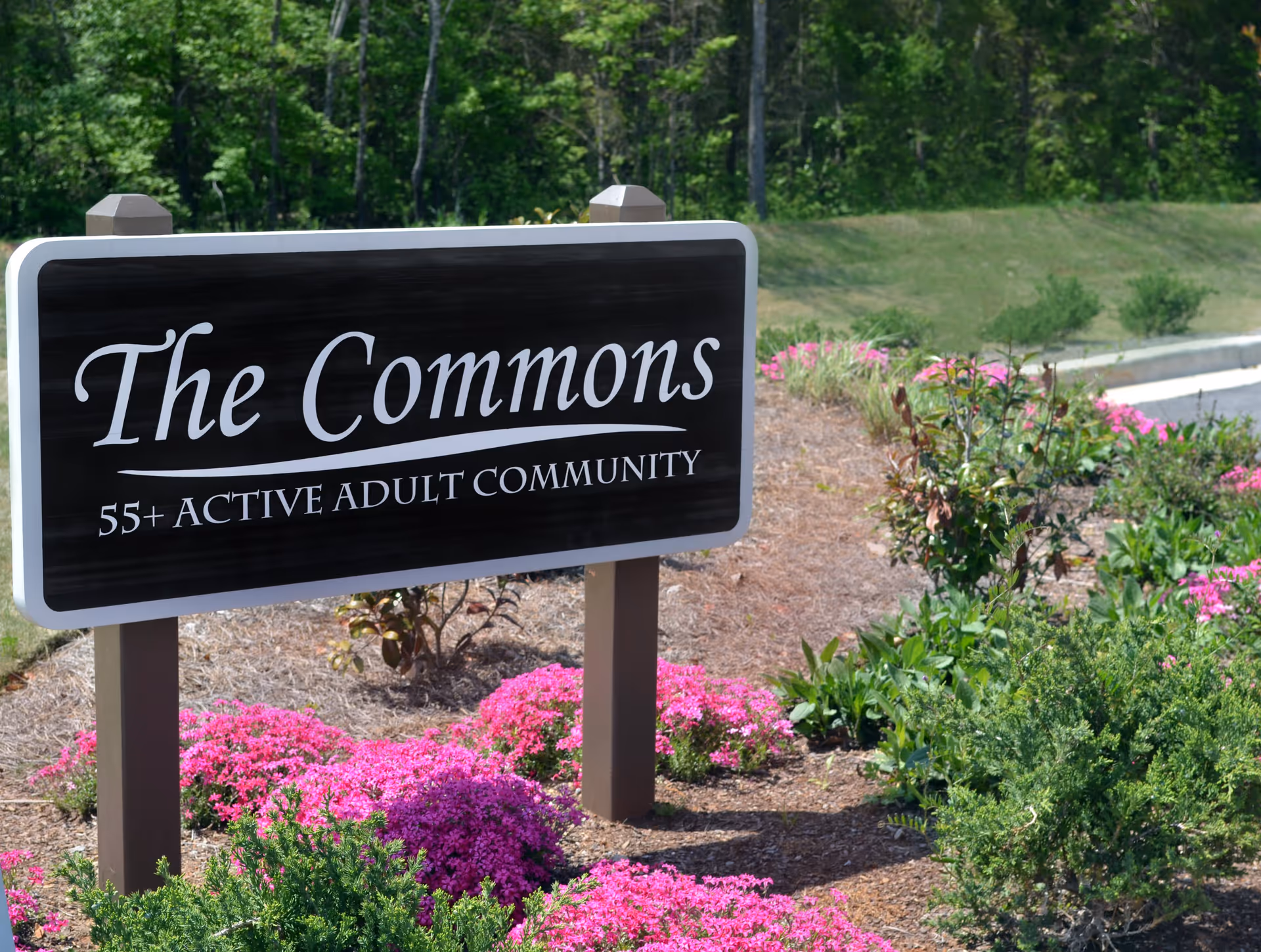 A black and white sign reading 'The Commons 55+ Active Adult Community' surrounded by green shrubs and pink flowers with a grassy area and trees in the background.