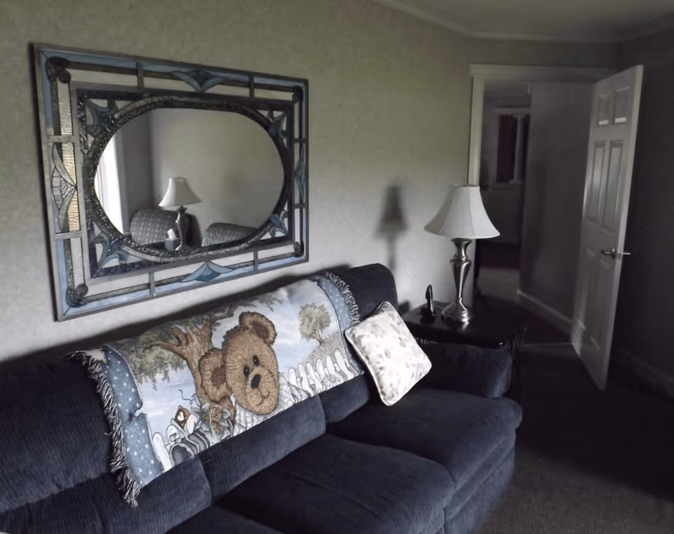 A dark upholstered sofa with a teddy-bear throw pillow beneath a decorative wall mirror and a side table with a lamp in a small living room.