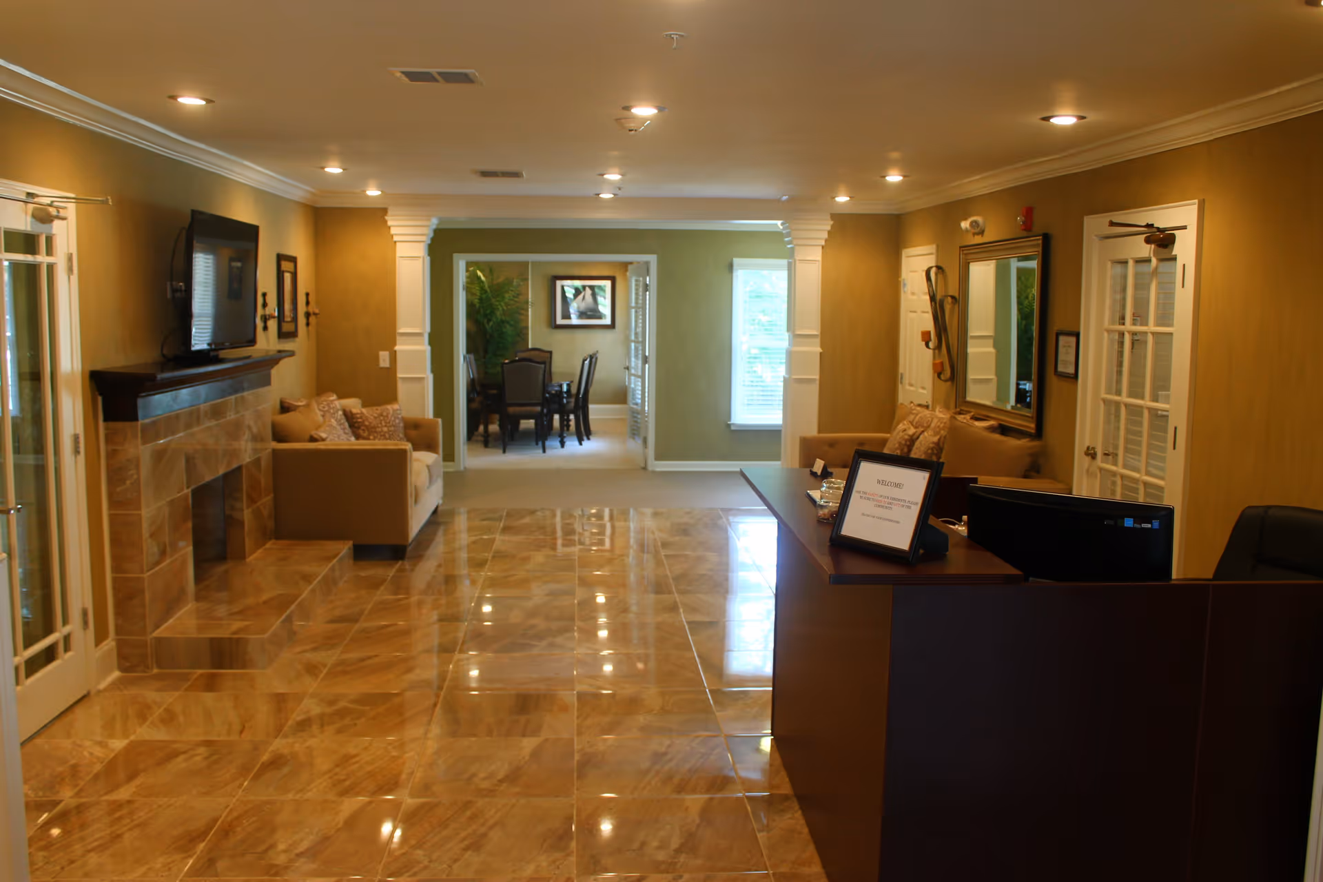 Interior view of a senior living facility reception area with a dark wooden reception desk, a computer monitor, and a framed welcome sign. The room features glossy tiled floors, a beige sofa with cushions, a wall-mounted TV above a tiled fireplace, and a dining area with a table and chairs visible through an open doorway. The walls are painted light brown, and there are decorative columns framing the entrance to the dining area.