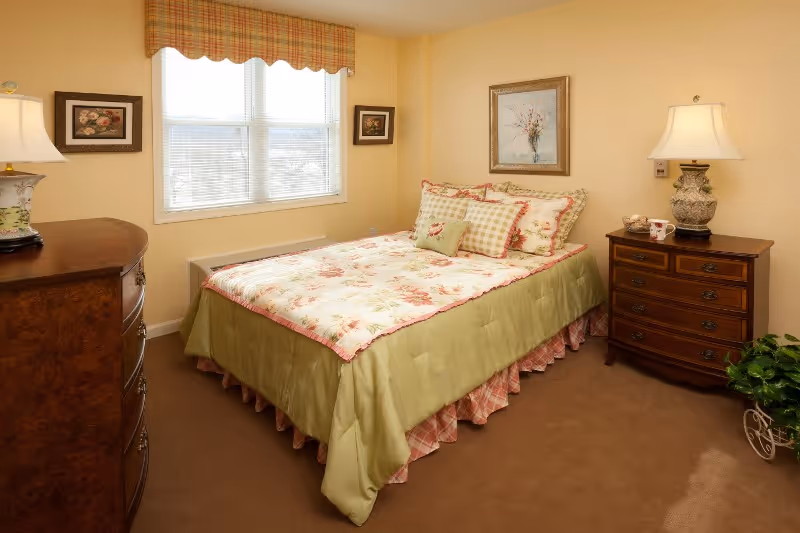 A cozy bedroom with a bed dressed in floral and checkered bedding, flanked by two wooden nightstands each with a lamp. There is a window with blinds and a valance, two framed pictures on the walls, and a small green plant in the corner.