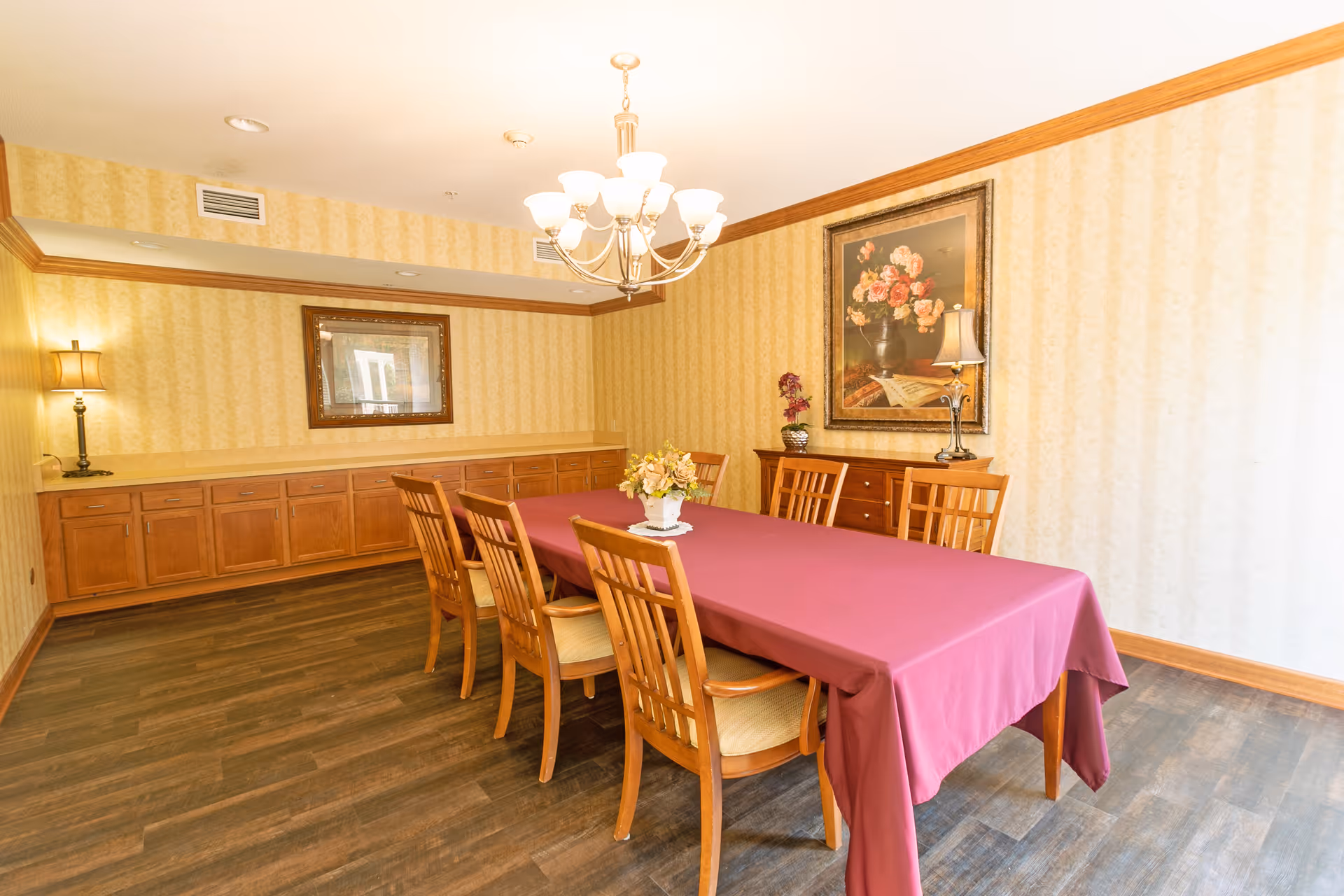 A dining room with a long rectangular table covered with a purple tablecloth and surrounded by eight wooden chairs with beige cushions. The room has wooden flooring, yellow patterned wallpaper, and wooden trim. There is a chandelier hanging above the table, a sideboard with a lamp and floral painting on the wall, and another lamp on a cabinet along the opposite wall.