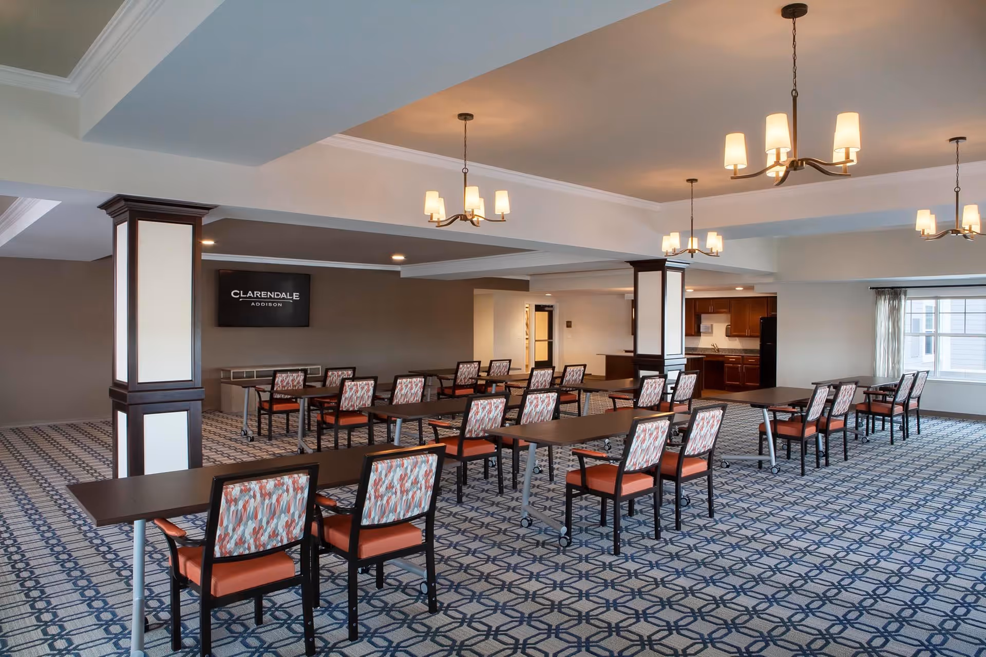 A spacious dining room with multiple rectangular tables and chairs arranged in rows. The chairs have patterned backs and orange seats. The room features patterned carpet flooring, several chandeliers with warm lighting, and large windows allowing natural light. A wall-mounted screen displays the text 'CLARENDALE ADDISON'.