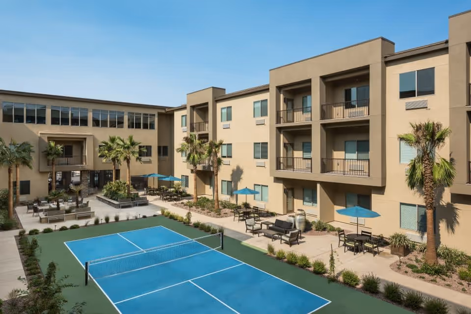 Outdoor courtyard area of Carnegie Heights at Henderson featuring a blue pickleball court surrounded by seating areas with tables, chairs, and umbrellas, palm trees, and a three-story beige building with balconies and windows under a clear blue sky.