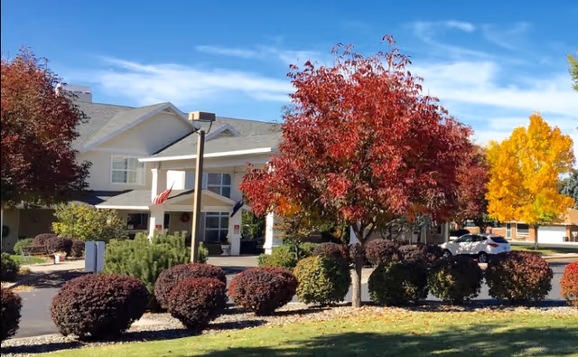 Exterior view of McKay Creek Assisted Living facility with a white building, a covered entrance, and a landscaped area featuring bushes and trees with autumn foliage under a clear blue sky.