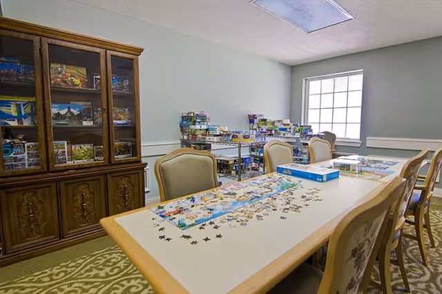 A well-lit room with a long table surrounded by chairs. On the table, there is a partially completed jigsaw puzzle with puzzle pieces scattered around. In the background, there is a wooden cabinet with glass doors displaying various items and shelves filled with board games. A window lets in natural light, brightening the room.