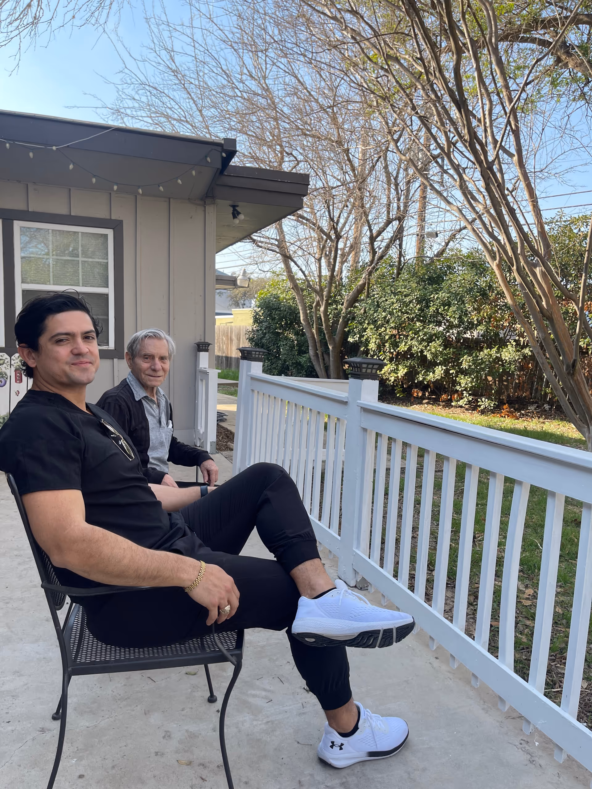 Two men sitting on metal chairs on a porch outside a building. One man is younger, wearing black clothes and white sneakers, and the other is older with gray hair, wearing a gray shirt and black jacket. Behind them is a white railing, trees without leaves, and green bushes under a clear blue sky.