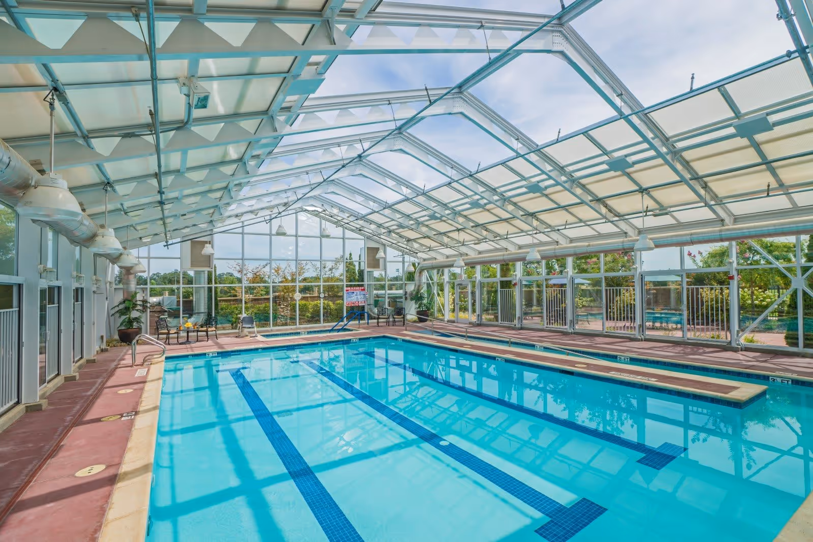 Indoor swimming pool with clear blue water under a glass roof structure, surrounded by glass walls showing outdoor greenery. There are chairs and tables along the poolside.