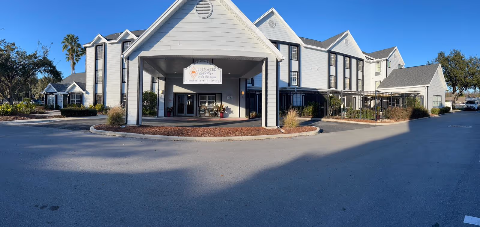 Exterior front view of Elevated Estates of New Port Richey, a multi-story senior living facility with white siding and black window frames. The entrance features a covered drop-off area with a sign displaying the facility's name and slogan, surrounded by landscaping and a clear blue sky.