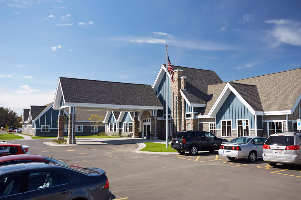 Exterior view of The Neighborhoods at Brookview facility showing a large building with blue and beige siding, multiple peaked roofs, a covered entrance, an American flag on a flagpole, and a parking lot with several cars parked.