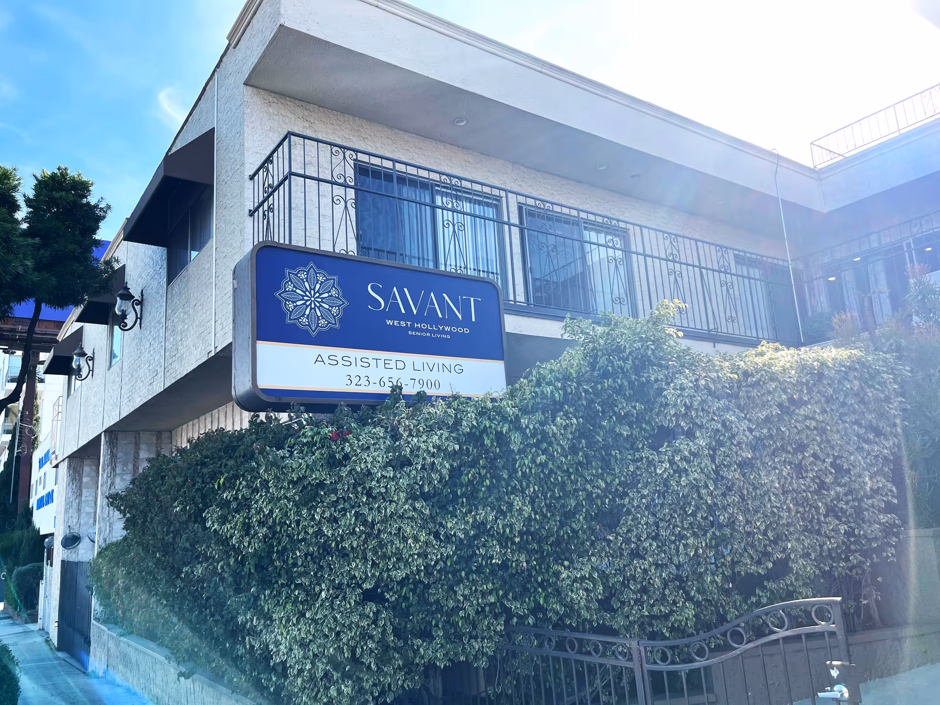 Exterior view of Savant of West Hollywood assisted living facility showing a two-story building with white textured walls, black wrought iron balcony railings, and a large blue and white sign with the facility name and phone number. There are green bushes in front of the building and a clear blue sky above.